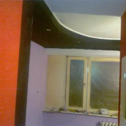 Wnętrze pokoju w trakcie prac wykończeniowych: ściany częściowo pomalowane na pomarańczowo i fioletowo, okno zabezpieczone folią, widoczny fragment sufitu podwieszanego z oświetleniem.